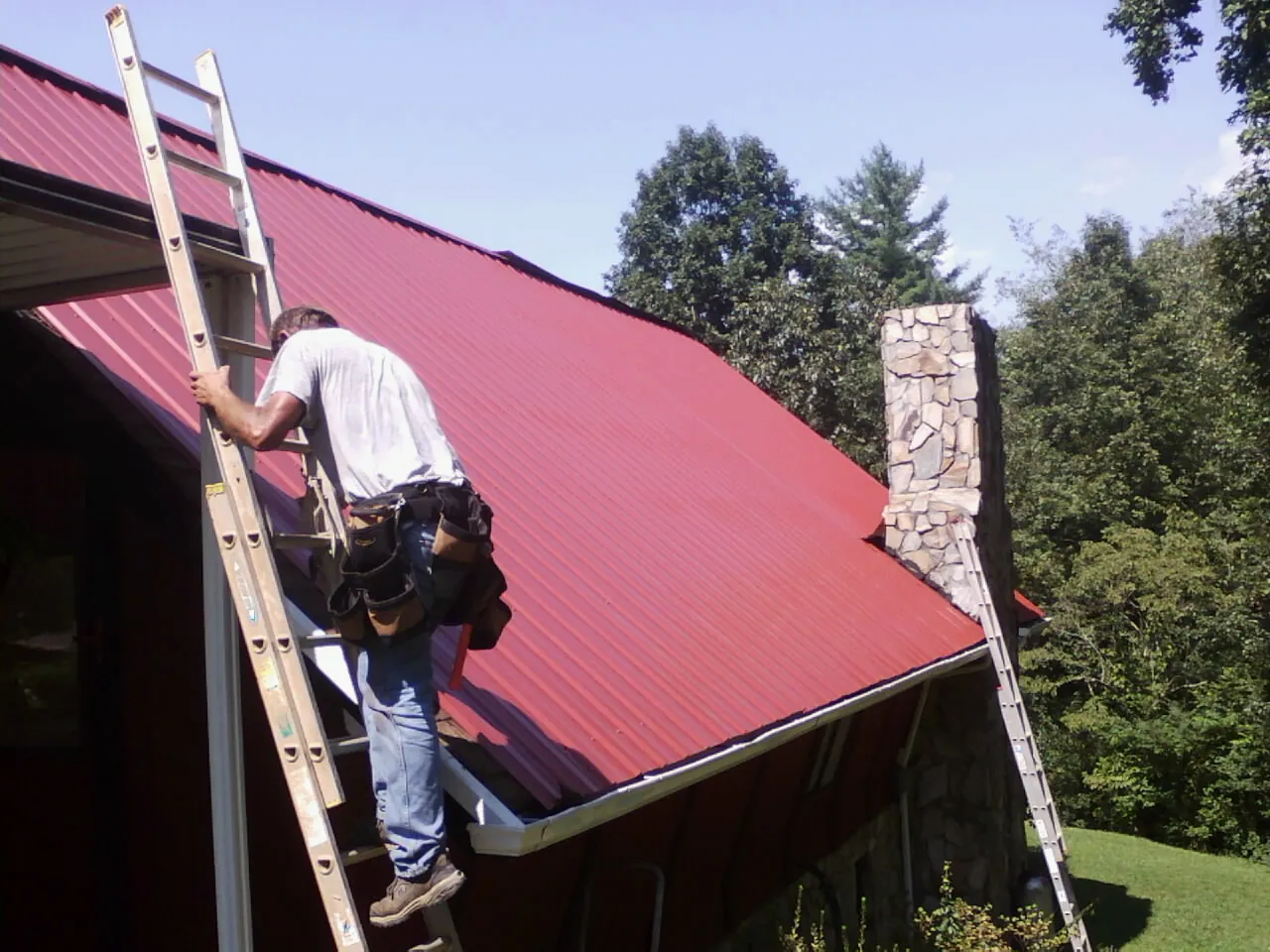 Roofer on a ladder beside a red metal roof and stone chimney — Soffit & Fascia Repair by Roofing Brothers in Mathiston